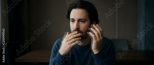 Concerned adult man, thinking deeply with hands near mouth, dark blue sweater and facial hair, dimly lit indoor home office interior, perfect for mental health awareness marketing and insurance.
