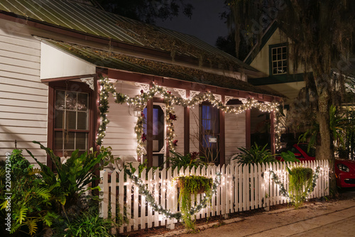 Glowing Christmas lights and garland decorate a small cottage in St. Augustine, Florida. A white picket fence surrounds the tropical garden and illuminated front porch at night.