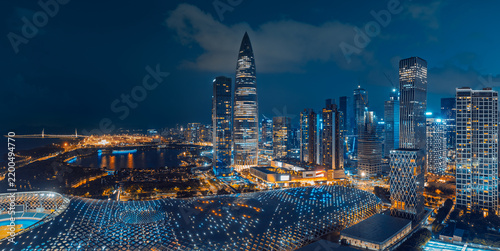 Illuminated Shenzhen skyline with a pointed tower, dense high-rises, waterfront park, bridges, and a curved glass roof beneath clouds at blue hour.