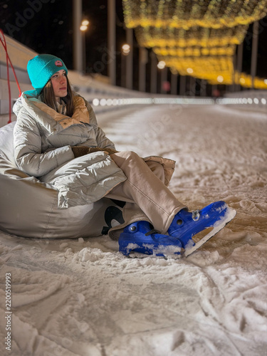 Woman sits on snow in winter while wearing colorful beanie and blue boots during nighttime near lights