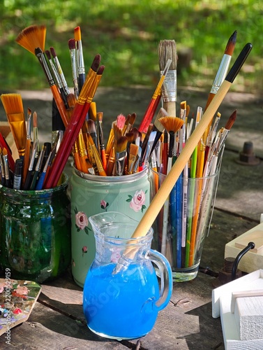 An assortment of painter tools with various art paintbrushes in glass jars and a pitcher with bright blue water on a rustic wooden table against a soft green background.