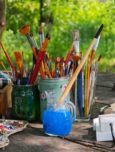 A creative outdoor painter artist workspace featuring multiple paintbrushes in jars, a glass jug filled with blue tinted water, and a wooden palette on a table in sunlight.