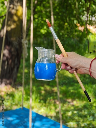 Woman holding a glass jar filled with vibrant blue paint and an artist paint brush in a sunny garden, creative outdoor painting activity concept and artistic leisure.