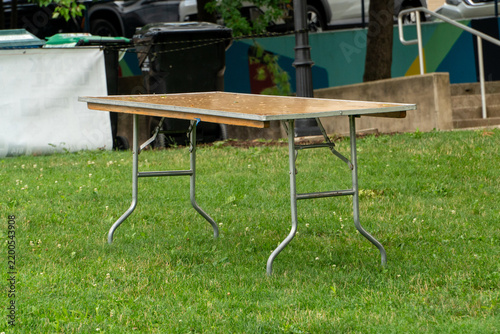 Фотография Empty portable folding table with wooden top and metal legs on vibrant green grass, ready for outdoor events, temporary setups, or community gatherings in a park setting