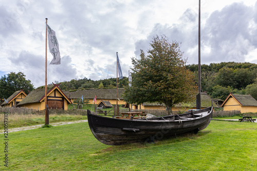 View of the Fyrkat Viking Museum featuring a traditional wooden Viking ship replica on a grassy field surrounded by historic thatched-roof longhouses in Hobro, Jutland, Denmark.