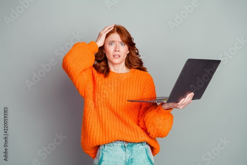 Foto Confused young redhead woman in orange sweater holding laptop and scratching her