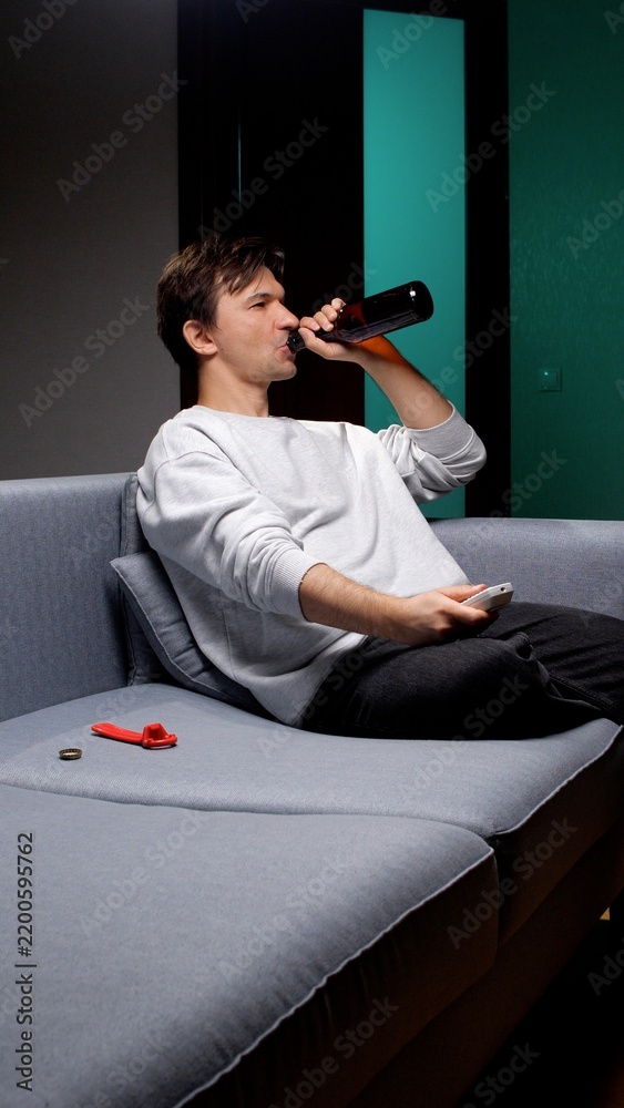 Fototapeta premium Young man lounging on a couch at home, holding a beer bottle and tv remote, relaxing in his living room and unwinding during an evening or weekend downtime
