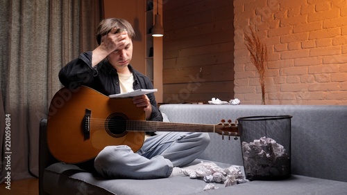 Obraz na plátně Young man on a sofa holding an acoustic guitar and crumpled paper, frustrated an