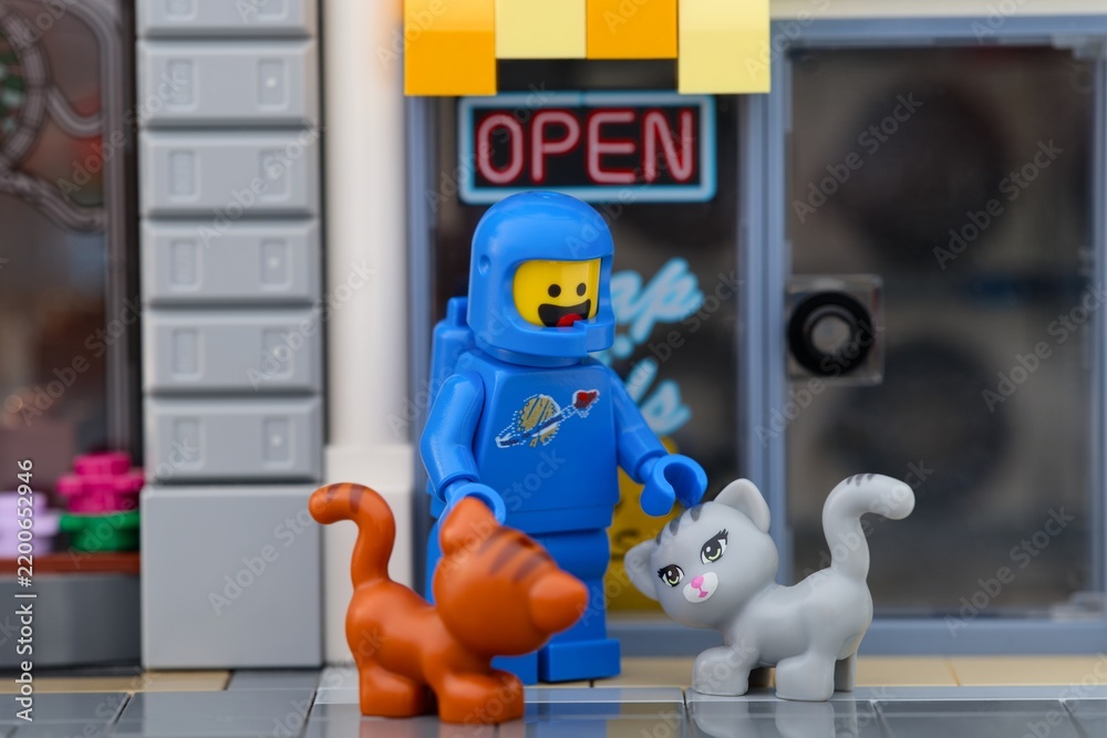 Obraz premium Tambov, Russian Federation - September 19, 2026 A blue Lego astronaut minifigure standing outside a shop with an Open sign, petting a grey cat while an orange cat standing nearby.