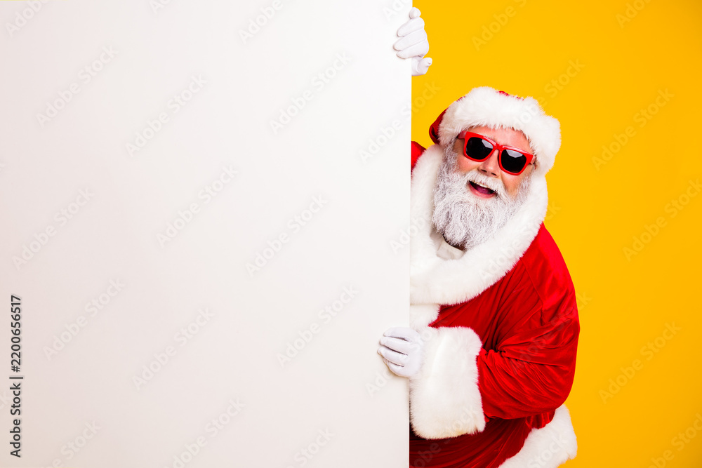 Obraz premium Santa Claus wearing red sunglasses peeking from behind a blank white sign on a yellow background — cheerful festive Christmas advertising and holiday greeting concept