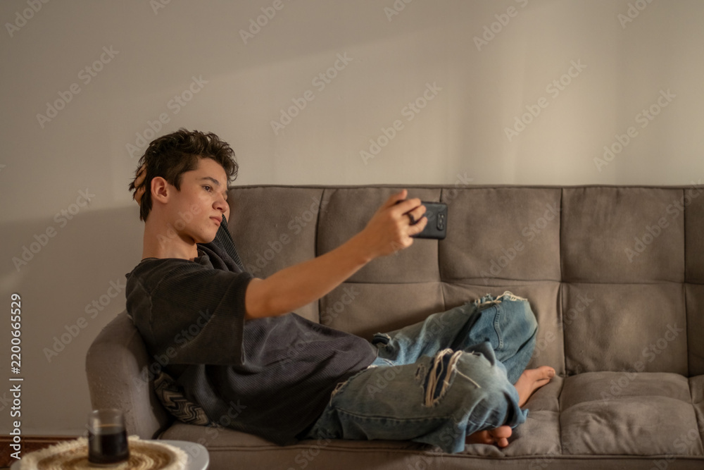 Fototapeta premium Teenager lies on sofa holding phone above himself for new shot, blogging repeated until frame feels right, emotion that stops scrolling. Wide shot. Concept of blogging habits of teenage boy