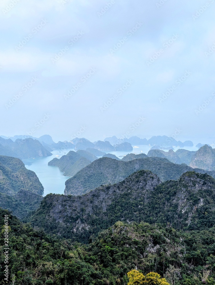 Fototapeta premium Elevated panoramic landscape showing sea waters winding between forested limestone karst peaks under a soft hazy blue sky in Lan Ha Bay.