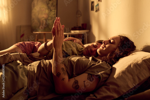 Young adult Caucasian romantic couple lying in bed resting together and holding hands at home. Image suitable for relationship wellness and lifestyle branding campaigns