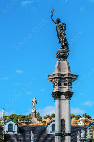 The centennial Monument to Independence in Quito in the foreground, with the Virgin of El Panecillo in the background, under a blue sky.