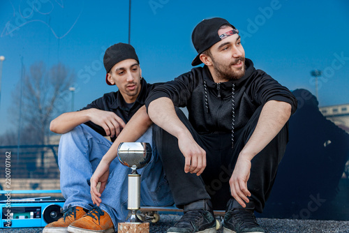 Foto Two young men sitting outdoors with a trophy, skateboard, and boombox, celebrati