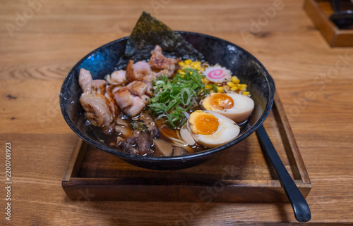 Bowl of ramen with sliced pork, soft-boiled eggs, green onions, and nori served on a wooden tray in a traditional Japanese restaurant setting.