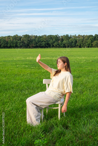 Young woman sitting alone on a white wooden chair in a lush green meadow.