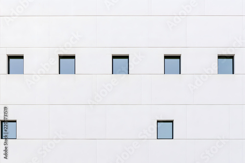 Minimalist white building facade with irregularly spaced square windows showing sky reflections. Clean, modern, geometric architecture.