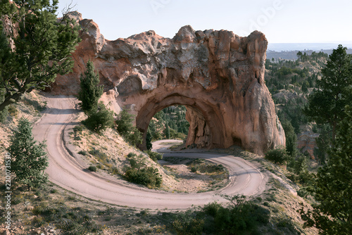 Scenic dirt road winding through a majestic natural rock arch in a mountainous, forested landscape under a clear sky.