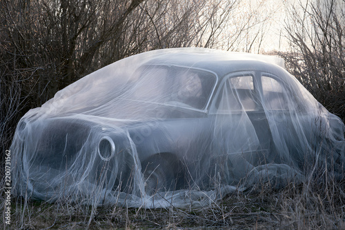 An old, rusty car covered with a sheer white fabric, abandoned in an overgrown field, evoking a sense of decay and mystery.
