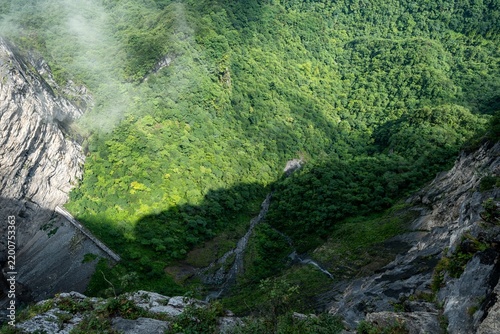 Bild auf Leinwand A dizzying downward shot revealing the sheer drop of a rocky mountain face, transitioning into deep, shadow-cast forests at the base of a wild canyon