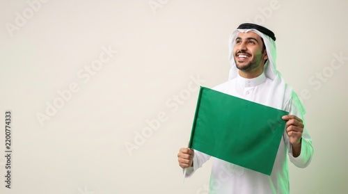 Smiling Saudi man holding national flag horizontally with both hands looking up against plain background   