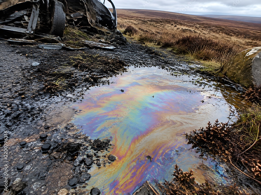 Obraz premium Iridescent diesel sheen pooling across a gritted moorland layby beside wreckage