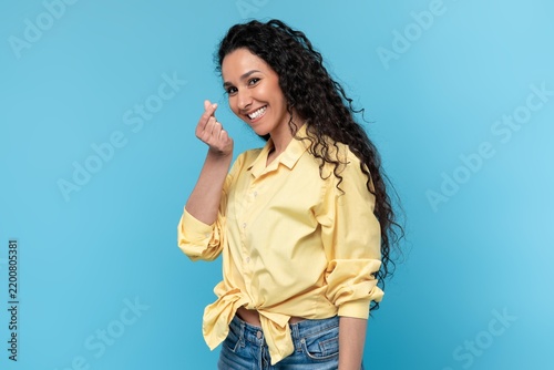 Cheerful Young Hispanic Woman Smiling And Gesturing With Hand Towards Camera, Standing In Studio Setting With Light Blue Background. Positive Emotion Displayed, Weight Loss Concept.