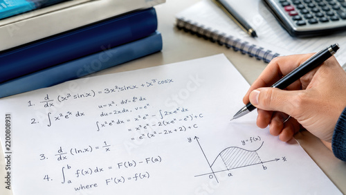 Close-up of a hand writing calculus notes with integrals, derivatives and a shaded area graph on a notebook beside books and a calculator