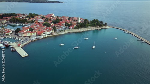 High wide drone shot flying over deep blue Adriatic waters revealing the historic harbor, stone breakwater, moored sailing boats, and red terracotta roofs of Novigrad Istria