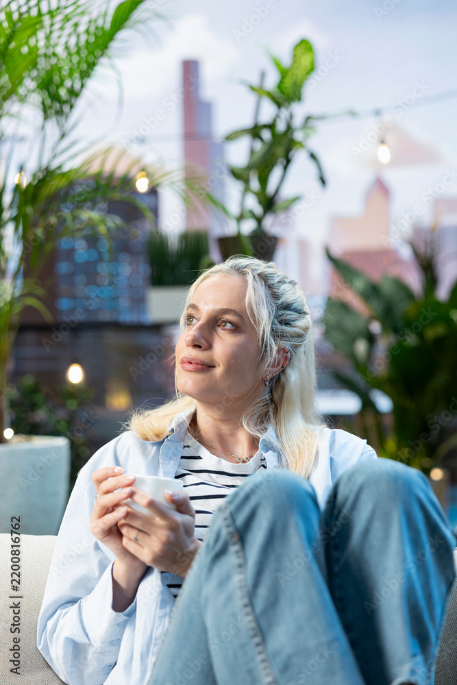 Fototapeta premium Relaxed woman holds a white coffee cup and looks upward while daydreaming on a rooftop sofa, enjoying a peaceful break in a modern urban outdoor lounge.