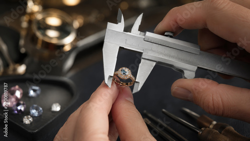 Close-up of hands measuring a diamond halo engagement ring with calipers on a jeweler's workbench