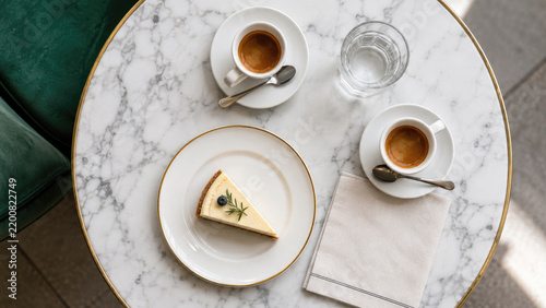 Top-down view of a marble café table with two espresso cups, a glass of water, a napkin and a slice of cheesecake garnished with blueberry and rosemary