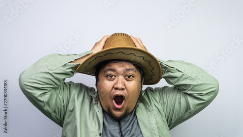 Foto Showcasing extreme surprise, this young Asian male grips his brown cowboy hat using both hands while standing before some plain light background
