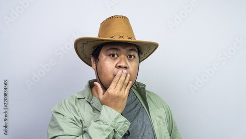 Obraz na płótnie Showcasing intense shock, a young Asian male covers his mouth using one hand while wearing western headwear plus green casual clothing before plain light background