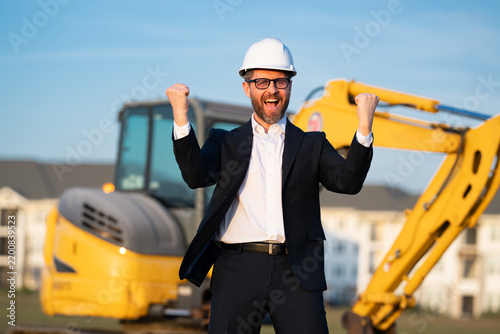 Construction owner near excavator. Confident construction owner in front of house. Architect, civil engineer. Man construction owner with a safety vest and hardhat at construction site.