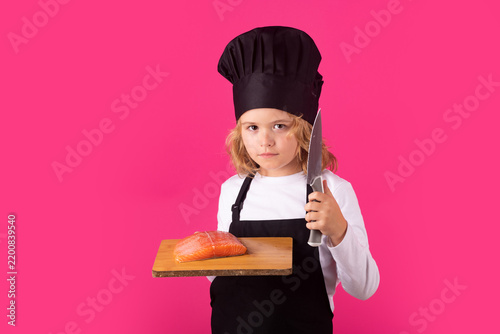 Kid cook hold cutting board with fish salmon steak and knife. Chef kid boy making healthy food. Portrait of little child in chef hat isolated on studio background. Child chef. Cooking process.
