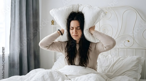 Stressed Young Woman Covering Ears with Pillow to Block Loud Noise in Bright Bedroom, Concept of Insomnia, Migraine, Sleep Disorder, and Morning Annoyance in Domestic Setting