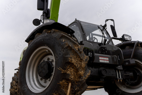 Low angle view of heavy agricultural telescopic handler with dirty tread tire, glass cabin, and lifting boom against overcast sky outdoors.