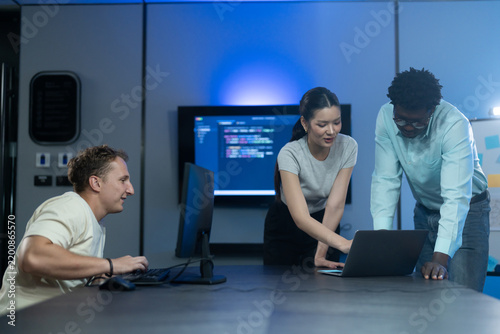 Asian female and African American male software developers discussing code on laptops with Caucasian colleague. Diverse tech team collaborating in modern IT office. Corporate technology, business data