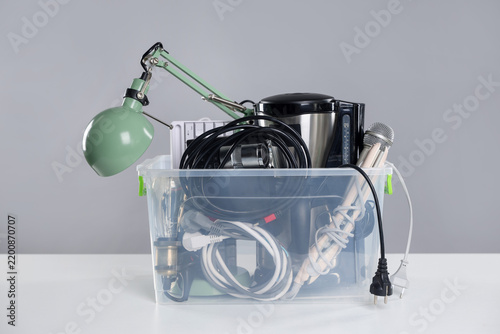 Plastic container with different broken household appliances for recycling on white table against light grey background