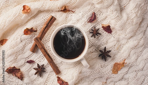 Top view of a steaming cup of hot coffee on a cozy white knitted sweater surrounded by cinnamon sticks, star anise, and autumn leaves.