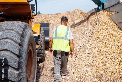 Worker in reflective vest walking beside heavy machinery and towering pile of wood chips at an outdoor industrial processing facility in daylight