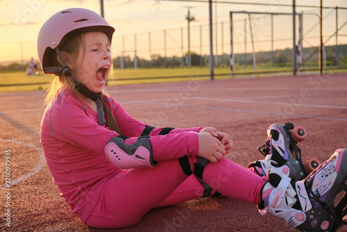 Young girl sitting on a sports field wearing a helmet and roller skates while showing a surprised expression