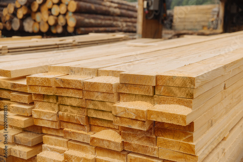 Stacked rough sawn timber planks with visible grain and cut ends arranged in a lumber yard with logs and boards in the background