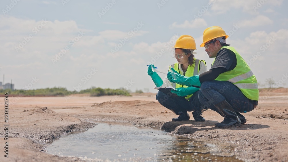 Obraz premium Asian male and female environmental inspectors kneeling by puddle to inspect water and soil pollution near factory. Environmental surveillance, ecological study and testing concept.