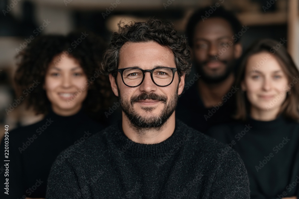 Fototapeta premium Smiling male team leader with glasses and beard stands in front of a diverse group of colleagues in a modern office portrait.