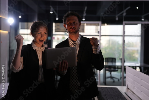 Fotografie An excited gasp turns into open cheering as the young woman's mouth widens in delight and her colleague raises both fists behind the laptop in the dark office interior
