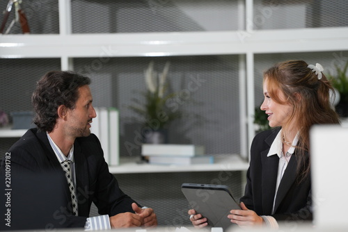 Fotografía Long-haired businessman in a patterned tie makes his case while the ginger-haired colleague angles her tablet and smiles by the white office shelving in a bright modern coworking office