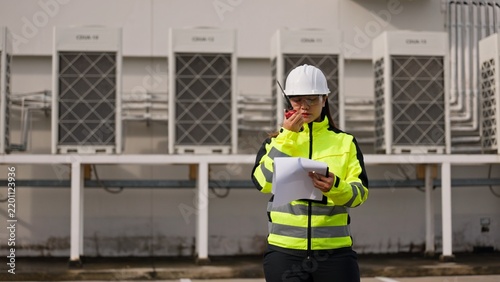 Asian female technician in hardhat checking equipment maintenance checklist on clipboard near cooling units. Commercial HVAC system service, facility engineering and safety concept.